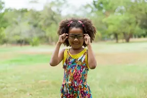 A smiling girl with curly hair is standing in a grassy field with her hands on her glasses.