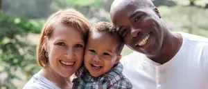 A family of three standing together and smiling for the camera in a park