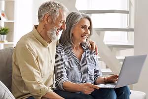 An older couple sitting on a couch, smiling, and looking at a laptop