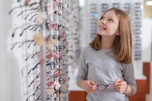 A girl with glasses standing in front of a wall with lots of eyeglasses displayed on it