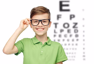 A young boy in a green polo shirt wearing glasses is smiling while holding up his glasses and looking at an eye chart in front of him.