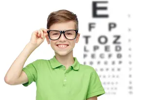A young boy in a green polo shirt wearing glasses is smiling while holding up his glasses and looking at an eye chart in front of him.