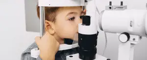 A young boy is looking into an eye examination machine in a clinic room.