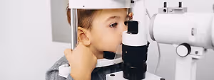 A young boy is looking into an eye examination machine in a clinic room.