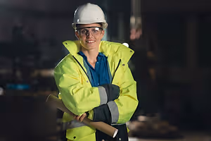 A woman in a yellow jacket and a white hard hat stands in a dimly lit room with a roll of paper in her left hand and a tool in her right hand.