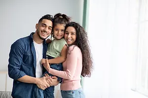 A family of three standing together and smiling for the camera, the woman is holding the child in her arms, and the man is holding the child's legs.