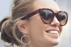 A smiling woman with brown hair is wearing sunglasses and earrings and is probably on a sunny day.