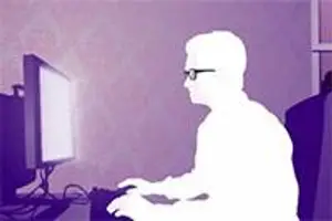A silhouette of a man wearing glasses is sitting in front of a computer monitor, with a slightly blurry background.