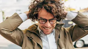A smiling man with curly hair wearing a tan coat and glasses stands outdoors in front of a building.