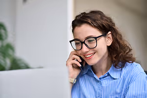 A woman wearing glasses and a blue shirt with white stripes is talking on her cell phone while sitting in front of a laptop on a desk.