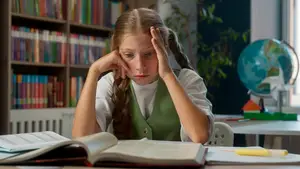 A girl in a green vest is sitting at a desk inside a classroom with her hands on her head.