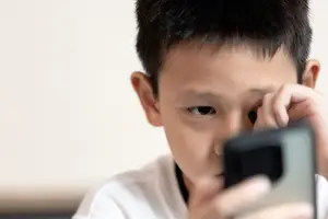A young boy with short black hair is holding a black cell phone with both hands and covering his face with his left hand.
