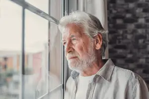 an elderly man with white hair looking out of a window with a building in the background