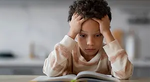 A boy with curly hair sitting at a table, holding his head with both hands and looking at an open book on the table.