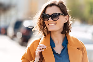 A smiling woman wearing sunglasses and a brown coat on a street