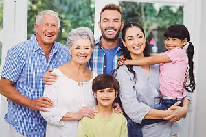 A smiling family of five, including an elderly man, a woman, a man, a boy, and a girl, posing for a photo in a room with a glass window and greenery outside