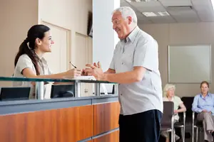 An elderly man speaking to a woman at a reception desk in a waiting room