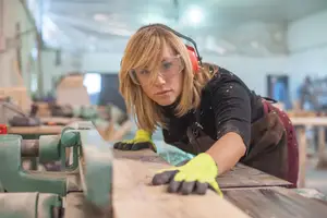 A woman in a workshop wearing goggles and gloves while cutting a piece of wood
