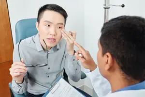 An Asian man sitting in a chair is talking to a doctor in a medical room.