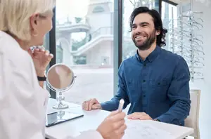 A man sits at a table in front of a woman with a mirror and tablet, possibly discussing eyeglasses