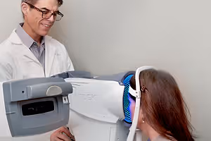 A man in a white lab coat and glasses is standing behind a woman who is using an eye examination machine with a blue light. The woman is smiling and looking at the machine, while the man is smiling and looking at her. They are standing in front of a white wall.