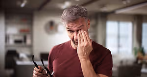 Image of an older man with gray hair wearing a maroon t-shirt holding glasses while rubbing his eye with his hand in an indoor area