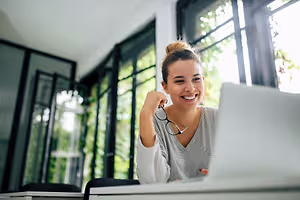A smiling woman is sitting in front of a desk with a laptop, holding a pair of glasses and smiling at the camera. Behind her, there is a glass wall with a view of the outside. On the left side, there is a glass window with a view of the trees. On the right side, there is a glass door with a view of the outside. On the right side, there is a glass window with a view of the trees.