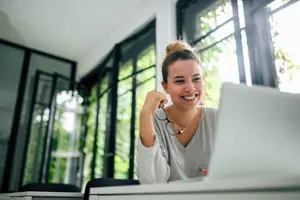 A smiling woman is sitting in front of a desk with a laptop, holding a pair of glasses and smiling at the camera. Behind her, there is a glass wall with a view of the outside. On the left side, there is a glass window with a view of the trees. On the right side, there is a glass door with a view of the outside. On the right side, there is a glass window with a view of the trees.