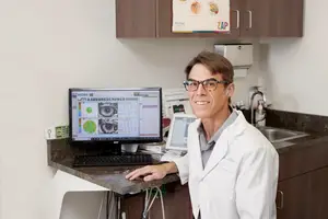 An adult male doctor in a white lab coat stands in front of a computer in an office