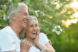 An older man and woman are laughing and posing for a picture in a garden with white flowers