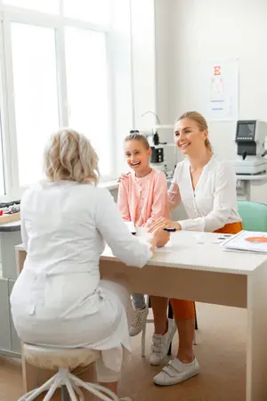 A woman doctor sits with a young girl and another woman in a room with a desk, a window, and an eye chart on the wall.