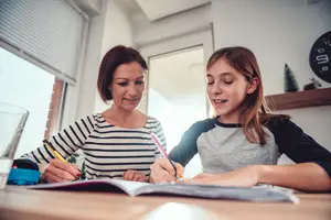 An adult woman and a young girl are sitting at a table and writing in a book together.