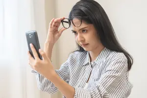 A woman wearing glasses is holding a phone and trying to focus on it, with a serious expression.
