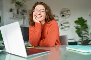 A woman is sitting in front of a desk, smiling and posing for a photo with a laptop, books, and a plant in the background.