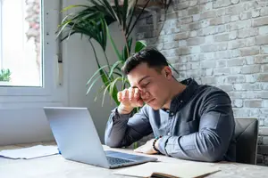 An adult man with short black hair sits with his eyes closed while holding his face with one hand in a room with a brick wall and a large window with a plant inside it
