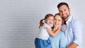 Image of a family with a young girl, a woman, and a man smiling at the camera against a white brick wall background.