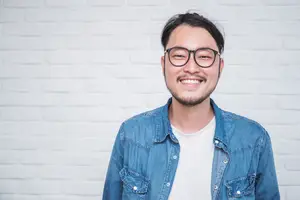 Man wearing a denim jacket and white t-shirt with a smiling face standing in front of a white brick wall