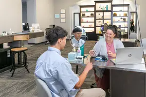 A man in a blue shirt is looking at himself in a mirror with a woman in a pink shirt and glasses smiling at him. Behind them is a desk with a laptop, and shelves with various items are in the background. A person is standing in the distance near a mirror. A wooden counter with a laptop and a bottle is on the left.