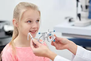 A young girl is holding a pair of glasses while smiling at the camera, with an optometrist checking her vision