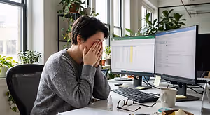 A woman sitting at a desk with computer monitors in front of her, looking stressed out