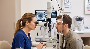A man is having his eyes examined by a female optometrist in a blue uniform using an eye examination device