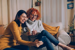 Two women sitting on a couch, one is holding a cup and a phone while the other one is smiling and looking at her phone