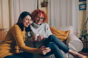 Two women sitting on a couch, one is holding a cup and a phone while the other one is smiling and looking at her phone