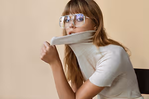 A woman sitting and adjusting her turtleneck with her left hand while wearing glasses.