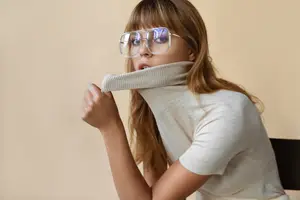 A woman sitting and adjusting her turtleneck with her left hand while wearing glasses.