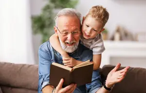 An elderly man is reading a book to a young boy on his lap on a couch.