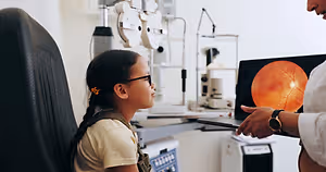 A girl with glasses is looking at an eye exam machine while a woman in a white coat holds a laptop showing an image of an eye.