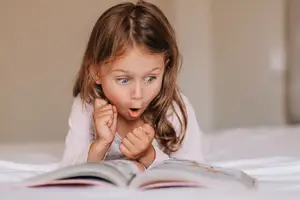 A little girl is lying on a bed and reading a book with her mouth open.