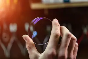 A hand holding a clear glass lens, possibly examining it, with a blurry background and a shelf with objects on it.