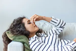 A woman in a striped shirt with a green blanket lying on a couch and blowing her nose with her hand